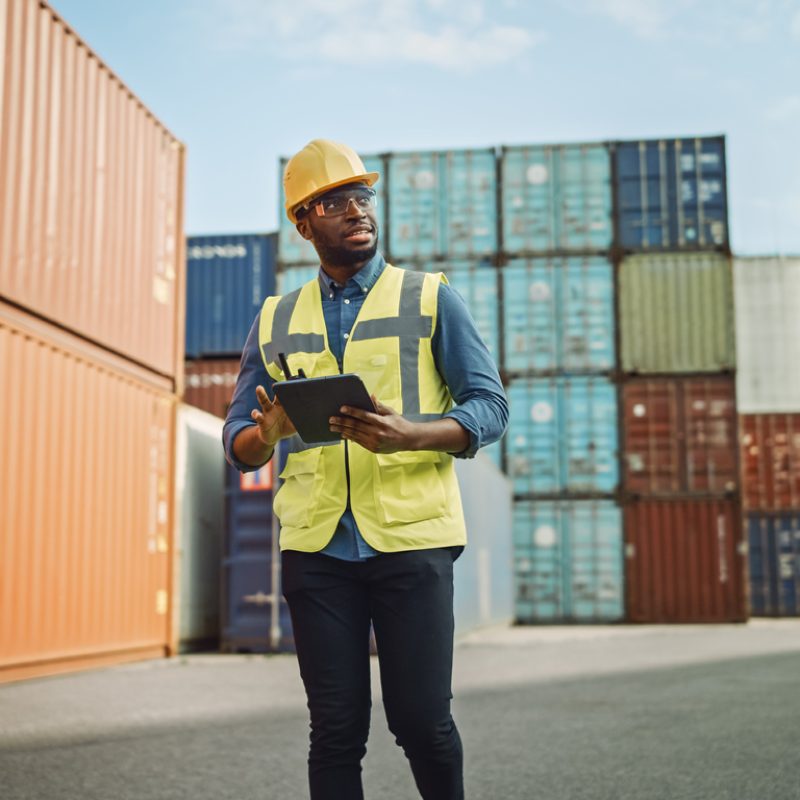 Smiling Handsome African American Black Industrial Engineer in Yellow Hard Hat and Safety Vest Working on Tablet Computer. Foreman or Supervisor in Container Terminal.